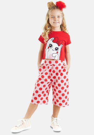 Camiseta roja corta con un gato de dibujos animados en blanco y un lazo rojo. Pantalones cortos rosas con grandes lunares rojos y bolsillos laterales funcionales. Zapatillas blancas.