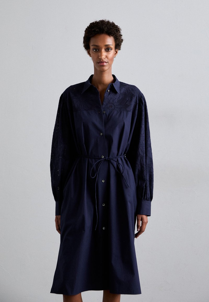 Granatowa sukienka zapinana na guziki z wiązaniem w talii, z długimi haftowanymi rękawami i usztywnionym kołnierzem. Gładka tekstura i długość do kolan.