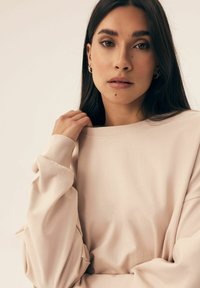 Jasnoróżowy, oversize'owy sweter z prążkowanymi mankietami i dekoltem, charakteryzujący się miękką fakturą materiału i luźnym krojem. Zbliżenie na górną część ciała.