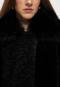 Melnā mētelī ar teksturētu vilnas audumu, gludu materiālu un pūkainu kažokādas apkakli. Zippera detaļa ir redzama priekšpusē.