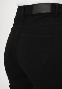 Zwarte high-waisted jeans met een gladde textuur, voorzien van twee achterzakken en een leren merklabel op de tailleband.