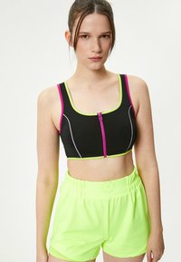 Sort sportsbh med pink og neongrøn detaljer, der har en lynlås foran. Sammen med neongrøn shorts, begge lavet af glat, strækbart materiale.