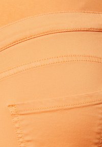 Persikofärgad tyg med en jämn textur, framträdande sömmar och en dold bakficka med kontrasterande sömdetaljer.