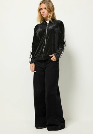 Blondynka ubrana w czarną welurową kurtkę na zamek z białymi paskami na rękawach oraz czarne szerokie spodnie, stojąca na jednolitym tle.