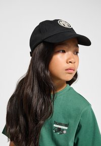 Svart baseballkeps med en rund broderad logotyp. Mjukt material. Satt ihop med en grön t-shirt med en logotryck på vänster bröst.