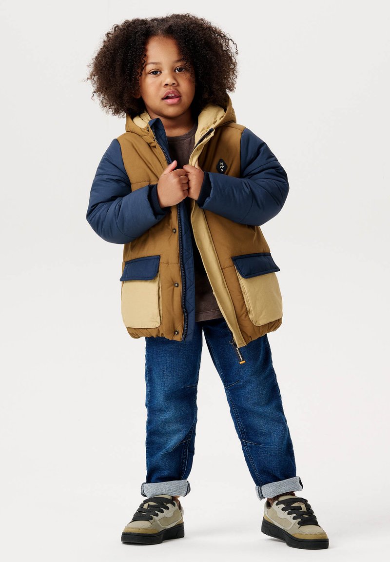 Puffjacke in einer Mischung aus Braun, Marineblau und Creme, mit Kapuze, zwei Fronttaschen und Reißverschluss. Kombiniert mit blauen Jeans und Sneakers.