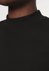 Svart ribbade polotröja med hög krage, med en slät textur och en figurnära passform. Närbild av nackområdet.