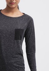 Langärmliges Oberteil aus dunkelgrauem meliertem Stoff mit einer glatten schwarzen Brusttasche und Raglanärmeln, das einen lockeren Halsausschnitt und eine weiche Textur aufweist.