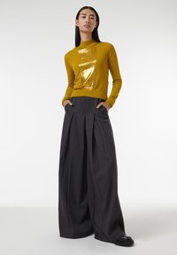 Senfgelbes Langarmoberteil mit einem Pailletten-Frontpanel; kombiniert mit hochgeschnittenen, weit geschnittenen, dunkelgrauen Hosen mit einem gefältelten Design.