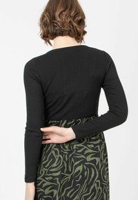 Schwarzes geripptes Langarmtop mit einem grünen Rock mit abstraktem Muster, der einen gerafften Bund und ein figurbetontes Oberteil hat.