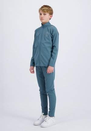 Boy standing in teal tracksuit and white sneakers, facing forward with neutral expression against plain light background.