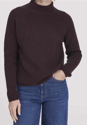Personne portant un pull en maille côtelée brun foncé et un jean bleu, une main dans la poche, sur un fond clair uni.