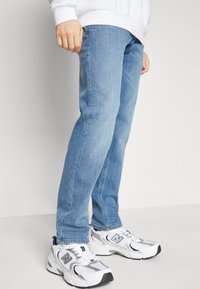 Person som bär ljusblå jeans och vita New Balance-sneakers, står mot en enfärgad vit bakgrund.