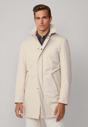 Hellbeige wasserdichte Jacke mit hohem Kragen, Druckknopfverschluss und zwei Seitentaschen. Glatte, matte Textur mit minimalen Akzenten.