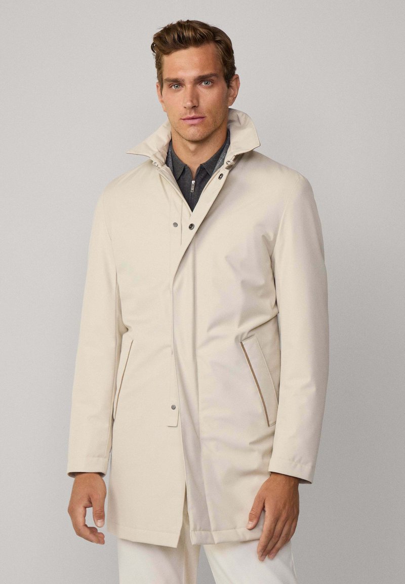 Chaqueta impermeable color beige claro con cuello alto, cierre a presión y dos bolsillos laterales. Textura suave y mate con acentos mínimos.