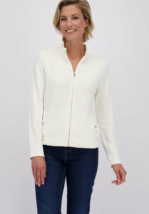 Vrouw die glimlacht, gekleed in een witte gestreepte ritsjas en donkerblauwe spijkerbroek, staande tegen een lichte effen achtergrond.