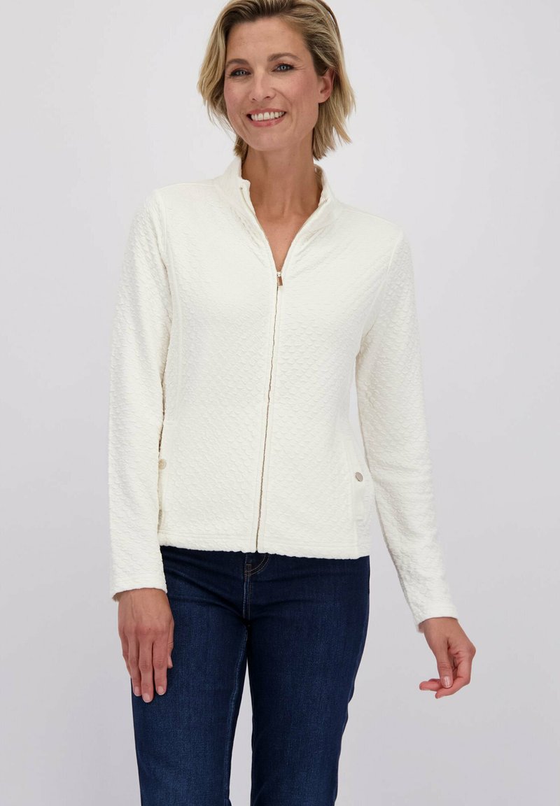 Femme souriante, portant une veste zippée blanche texturée et un jean bleu foncé, se tenant devant un fond clair uniforme.