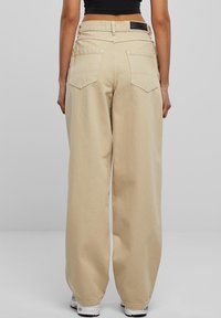 Béžové kalhoty s širokými nohavicemi a vysokým pasem, vyrobené z bavlny, se dvěma zadními kapsami a černou logistickou cedulkou. Hladká textura.