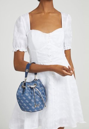 Kobieta w białej sukience zapinanej na guziki, trzymająca na ramieniu niebieską torebkę typu kubełek z krótkim paskiem.