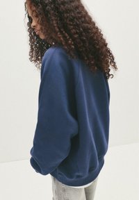 Felpa oversized blu navy con polsini e orlo ribattuti, caratterizzata da una texture liscia e un design minimalista. Indossata con jeans grigio chiaro.
