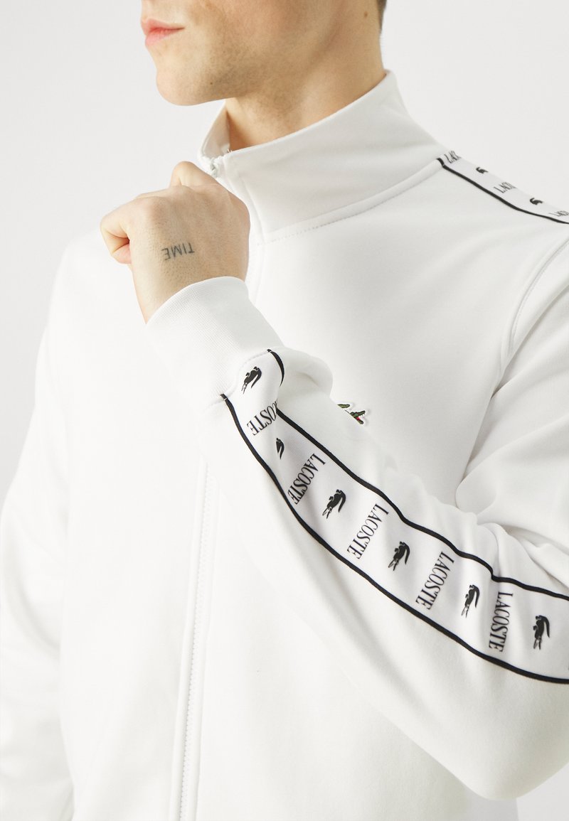 Witte ritssluitingjack met hoge kraag, voorzien van zwarte gestreepte accenten en het Lacoste-logo. Glad materiaal met een getailleerd ontwerp.