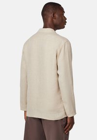 Beige Leinenhemd mit einfachem Kragen, langen Ärmeln und leicht abgerundetem Saum. Weiche Textur, lässige Passform und minimale Details.