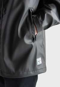 Chaqueta impermeable negra con una textura suave. Presenta un bolsillo lateral y una etiqueta blanca de Herschel cerca del dobladillo. Primer plano de la manga y el bolsillo.