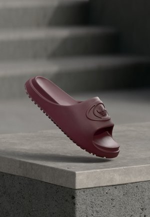 Dunkelrote Adidas Slide-Sandale mit strukturierter Sohle und Logo am Riemen, balanciert auf einem grauen Betonkante mit unscharfen Treppen im Hintergrund.
