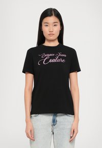 Juodas marškinėliai su rožine, kursyvine siuvinėjimo etikete "Versace Jeans Couture". Trumpomis rankovėmis ir apvaliu iškirpimu. Nešiojami su šviesiai mėlynomis džinsinėmis kelnėmis.