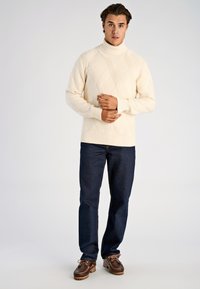 Crème trui met een getextureerd dambordpatroon, hoge halslijn en geribbelde manchetten, gecombineerd met donkerblauwe jeans en bruine schoenen.