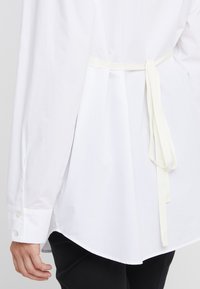 Vit långärmad skjorta med rundad nederkant, med ett mjukt krämfärgat bälte vid midjan och knäppte manschetter. Slätt bomullsmaterial.