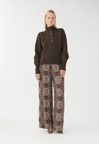 Brauner gerippter Strickpullover mit hohem Kragen und einer Knopfleiste mit fünf Knöpfen, kombiniert mit gemusterten, weiten Hosen in Braun- und Cremetönen.