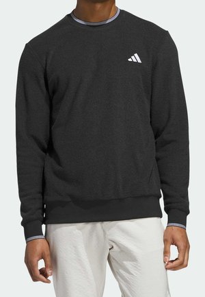 Mann trägt schwarzen Langarm-Sweatshirt mit kleinem weißem Adidas-Logo auf der Brust und hellen Hosen.