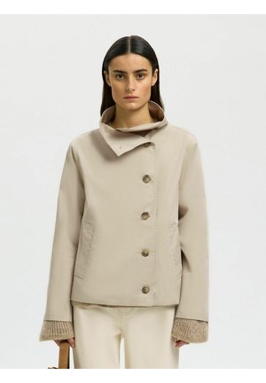 Mujer con cabello lacio y oscuro que lleva una chaqueta beige abotonada sobre un suéter de punto, sosteniendo un bolso marrón claro, frente a un fondo liso y claro.