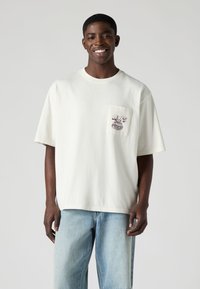 Άσπρο βαμβακερό t-shirt με άνετη εφαρμογή, κοντά μανίκια και τσέπη με μοβ γραφικό σχέδιο. Συνδυασμένο με ανοιχτόχρωμο μπλε τζιν.