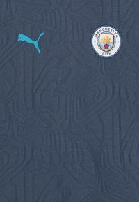 Temno modra športna majica z teksturiranem vzorcem. Ima svetlomodro Puma logo na levem prsnem predelu in krožni grb Manchester City, ki je centriran.