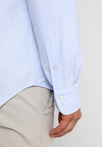 Lichtblauw overhemd met lange mouwen en knoopmanchetten, gemaakt van een gladde stof. Heeft een ontspannen pasvorm en een subtiele textuur, gecombineerd met beige broek.