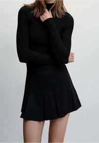 Donna che indossa un top a maniche lunghe nero a coste e una mini gonna nera plissettata, con le braccia incrociate e una mano che tocca il collo.