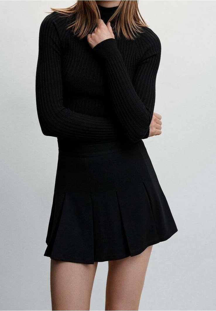 Donna che indossa un top a maniche lunghe nero a coste e una mini gonna nera plissettata, con le braccia incrociate e una mano che tocca il collo.