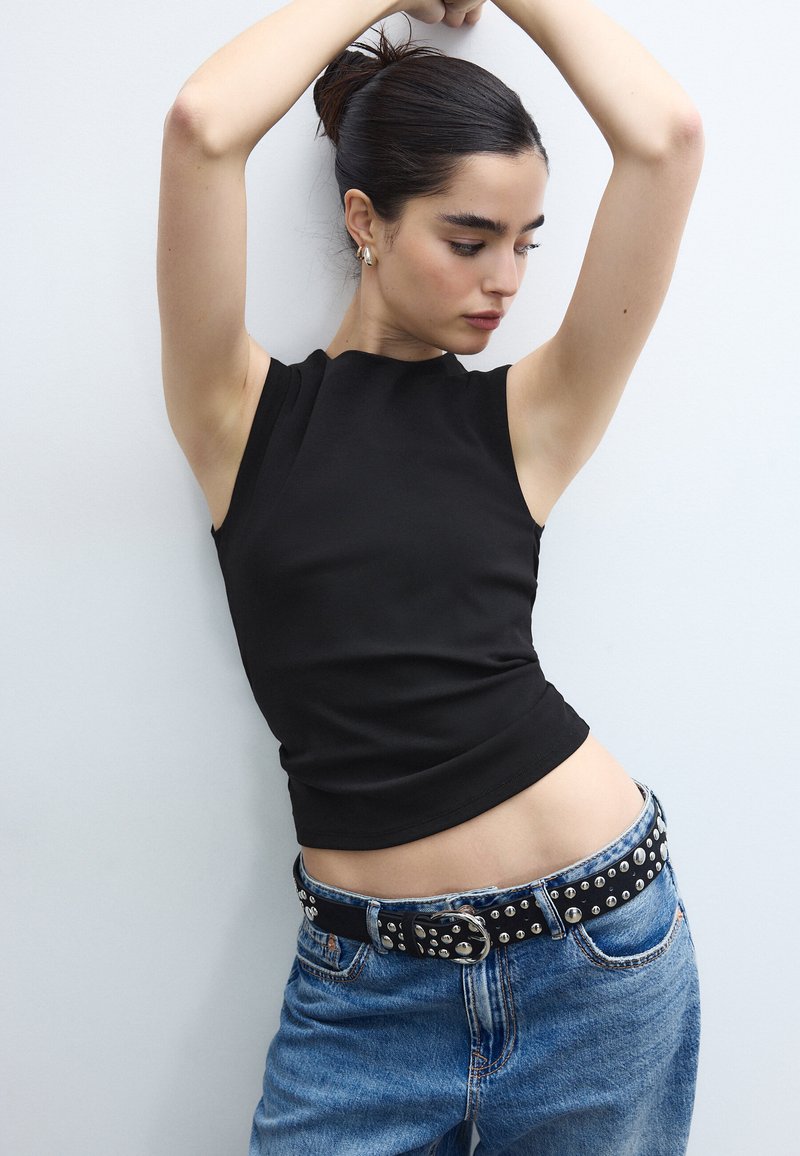 Frau mit dunklem Haar zu einem Dutt gebunden, trägt ein ärmelloses schwarzes Oberteil und einen mit Nieten besetzten Gürtel zu blauer Jeans, posiert vor einem schlichten weißen Hintergrund.