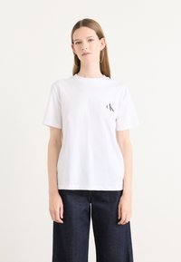 White cotton t-shirt with a round neck, short sleeves, and a small black logo on the left chest. Paired with dark blue wide-leg jeans.