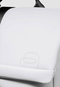 Witte tas met een matte afwerking, voorzien van een zwarte riem, een gladde textuur en een subtiel reliëflogo. Rechthoekige vorm met afgeronde hoeken.