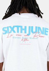 Witte katoenen T-shirt met grote blauwe tekst "SIXTH JUNE" en roze accenten met zinnen in een grafisch ontwerp op de achterkant. Simpele ronde hals.