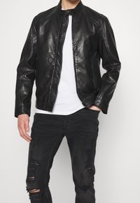 Zwarte leren jas met een staande kraag, ritssluiting en subtiele stikdetails. Draagt boven een wit t-shirt en gecombineerd met versleten zwarte jeans.