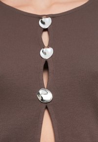 Καφέ ριγωτό τοπ με άνοιγμα σε σχήμα κλειδιού και τρία γυαλιστερά, ακανόνιστα ασημένια κουμπιά μπροστά. Ομαλή υφή και εφαρμοστή γραμμή.