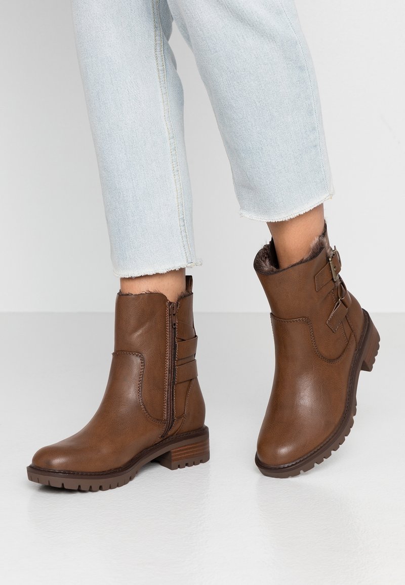 Botas de tobillo marrones con forro de pelo, cremallera lateral, correas con doble hebilla, combinadas con unos pantalones vaqueros cortos de mezclilla azul claro sobre un suelo blanco.
