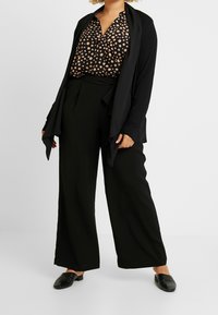 Cardigan lungo nero su una blusa a pois neri e beige, abbinato a pantaloni svasati neri e scarpe slip-on nere. Texture del tessuto liscia.