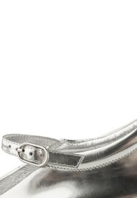 Zapato plateado metálico con punta redonda, que presenta una delgada correa en el tobillo asegurada por una pequeña hebilla de metal y un interior texturizado.