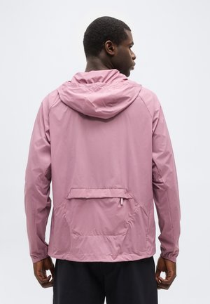 Moški, obrnjen stran, oblečen v svetlo roza kapucar s torbico na hrbtu in črne hlače, na enobarvni svetli podlagi.