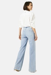 Vrouw die glimlacht, met haar rug naar de camera, gekleed in een wit longsleeve shirt en lichtblauwe wijdvallende jeans met hakken.